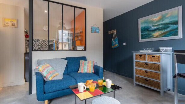 Ein Wohnzimmer mit einem blauen Sofa, bunten Kissen, einem Glastisch mit Frühstücksgebäck, einem Korbmöbelregal und einem großen abstrakten Gemälde an der Wand.