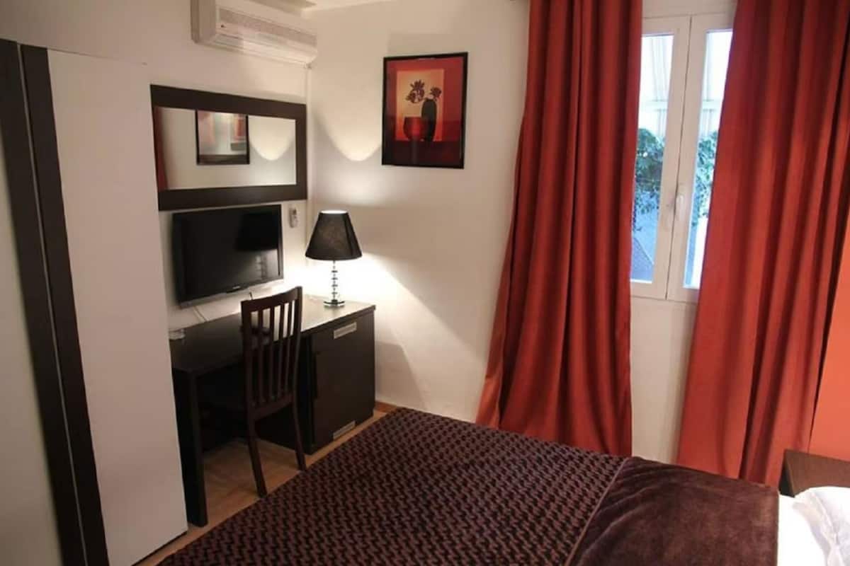 A hotel room with a bed, a desk with a chair, a TV, and a window with red curtains.