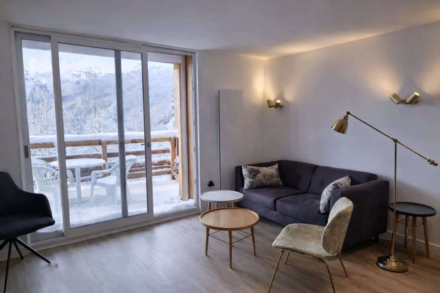 Un salon moderne doté d’une grande porte-fenêtre coulissante, d’un canapé gris foncé, d’une table basse ronde en bois, d’un fauteuil à motifs et d’un lampadaire.