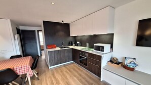 A modern kitchen with a central island, a dining area with a checkered tablecloth, and a small side table with magazines. - Beautiful, spacious apt near Arc 1600 (Bourg-Saint-Maurice)