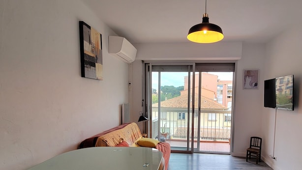 A modern living room with a glass-top dining table, a sofa, and a large sliding glass door leading to an outdoor balcony. - Charming love nest near the sea (Fréjus)