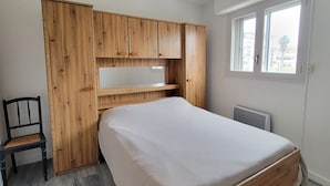 A bedroom with a wooden bed frame, a chair, a built-in desk, and a window with a view of buildings. - Charming love nest near the sea (Fréjus)