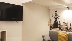 A living room with a grey sofa, a yellow pillow, a flat-screen TV, and a wall-mounted lamp.