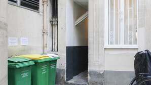 A narrow alleyway with a bicycle parked on the right, a window with decorative bars, and two green waste bins on the left.
