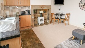 A compact kitchen with wooden cabinets, a stone fireplace, a dining area with a table and chairs, and a clock on the wall. - Luna Cottage (Holmfirth)
