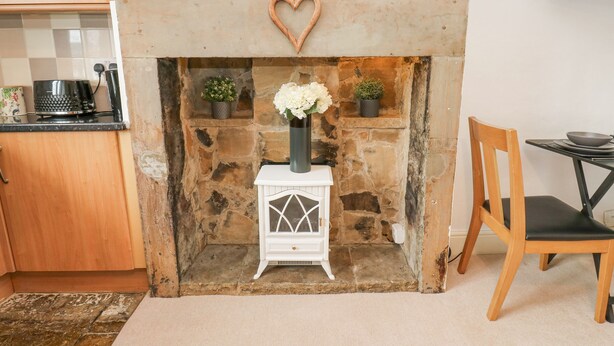 A cozy kitchen with a stone fireplace, a white decorative cabinet, and a dining area with a wooden chair and table. - Luna Cottage (Holmfirth)