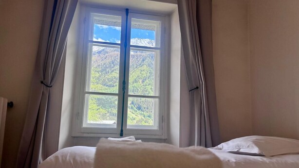 A bedroom with a large window, a bed with white linens, and a view of a forested mountain range. - Lovely flat in the heart of Chamonix (Chamonix-Mont-Blanc)
