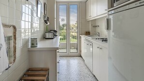 A narrow kitchen with white cabinets, a microwave, and a refrigerator. There is a small wooden stool and a window with a view of greenery outside. - In the heart of the Cap d'Antibes lovely flat (Antibes)