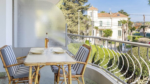 A balcony with a table and chairs, overlooking a well-maintained garden and a white building with a red roof. - In the heart of the Cap d'Antibes lovely flat (Antibes)