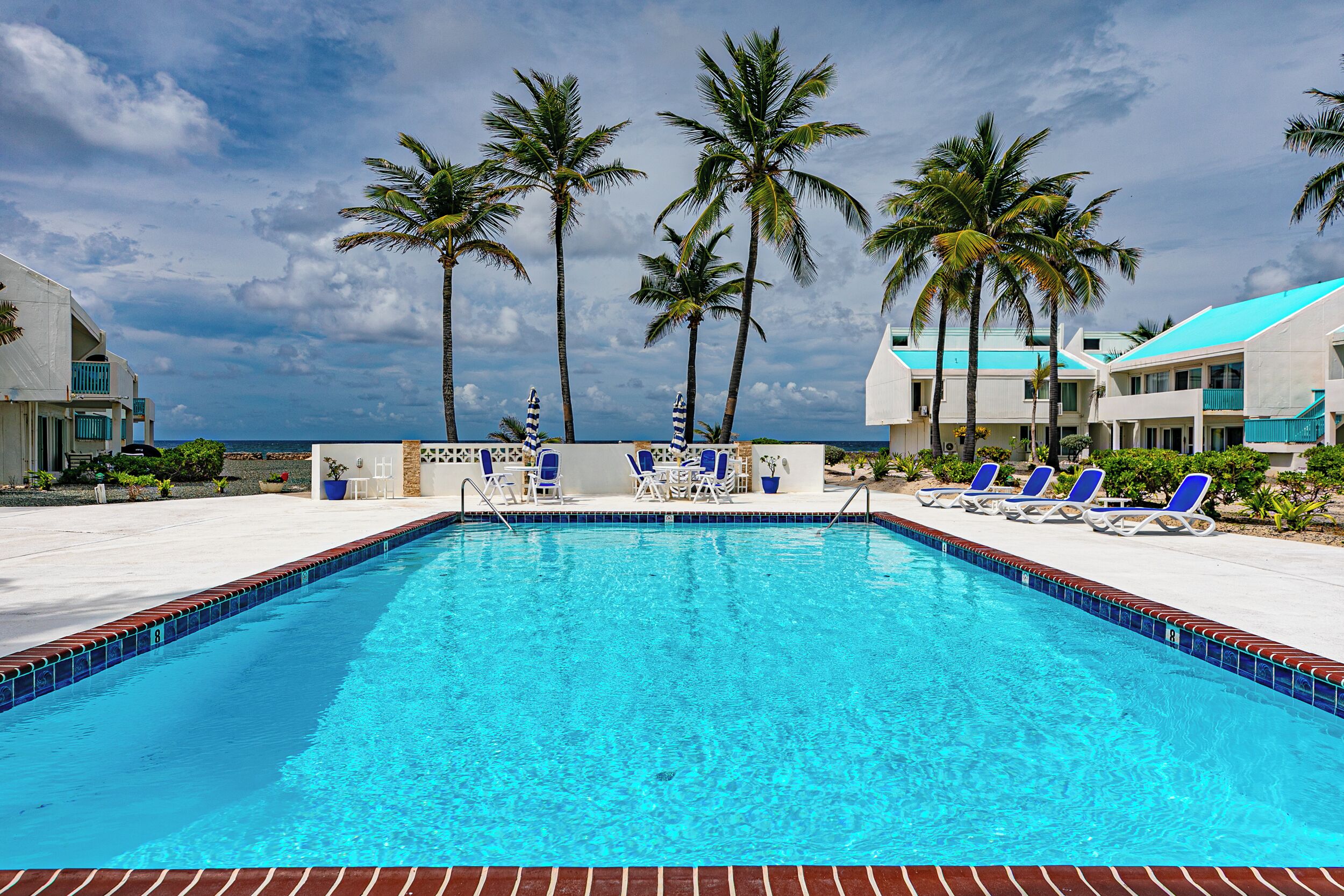 Ein Swimmingpool mit Liegestühlen und Palmen, umgeben von Gebäuden mit Blick auf den Ozean.