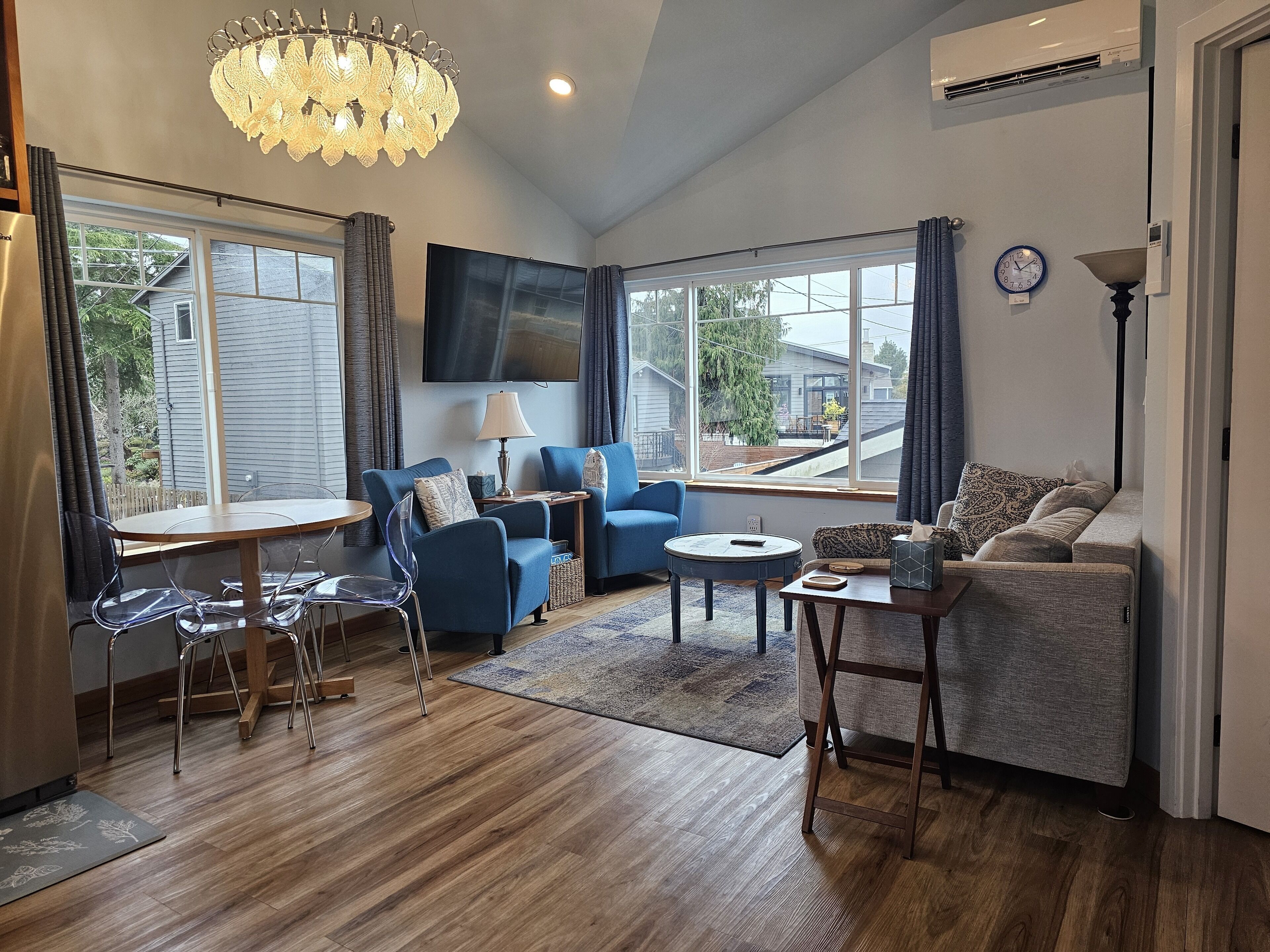 A modern living room with a dining area, large windows, a chandelier, and a flat-screen TV.