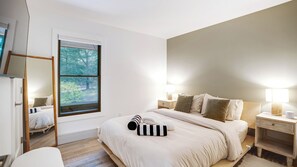 A bedroom with a large window, a bed with white bedding, two bedside tables with lamps, and a wooden nightstand. - Newly Renovated, Serene Property with Heated Pool, Tennis, and Resort-Like Spa (Southampton)