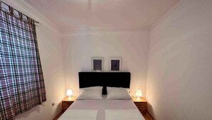 A bedroom with a bed, two bedside lamps, a headboard, and a window with a checkered curtain. - Holiday apartment with Wi-Fi (Novi Vinodolski)