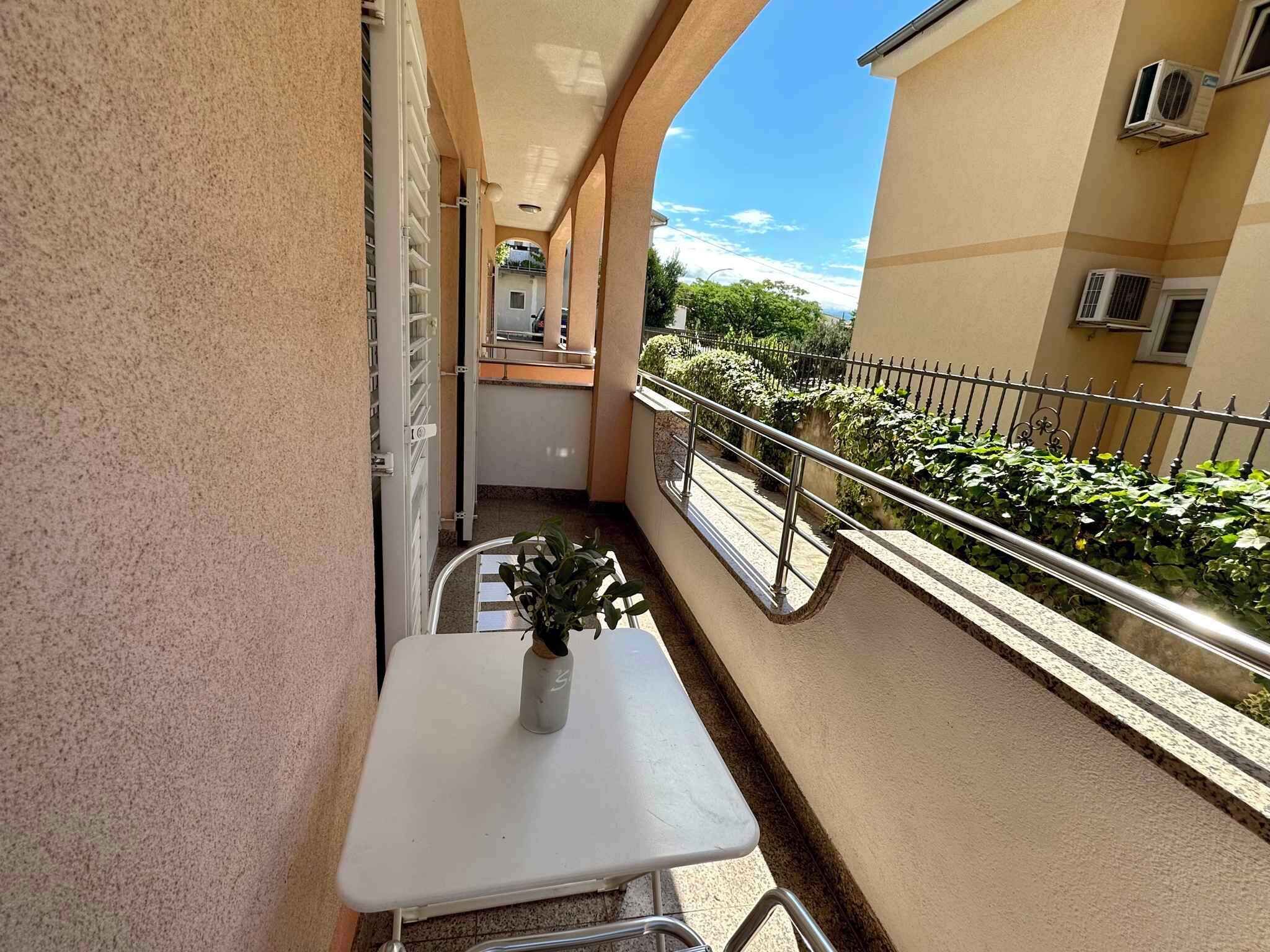 Un balcon avec une table blanche et un pot de fleurs, une balustrade et une vue sur un immeuble équipé de climatiseurs.