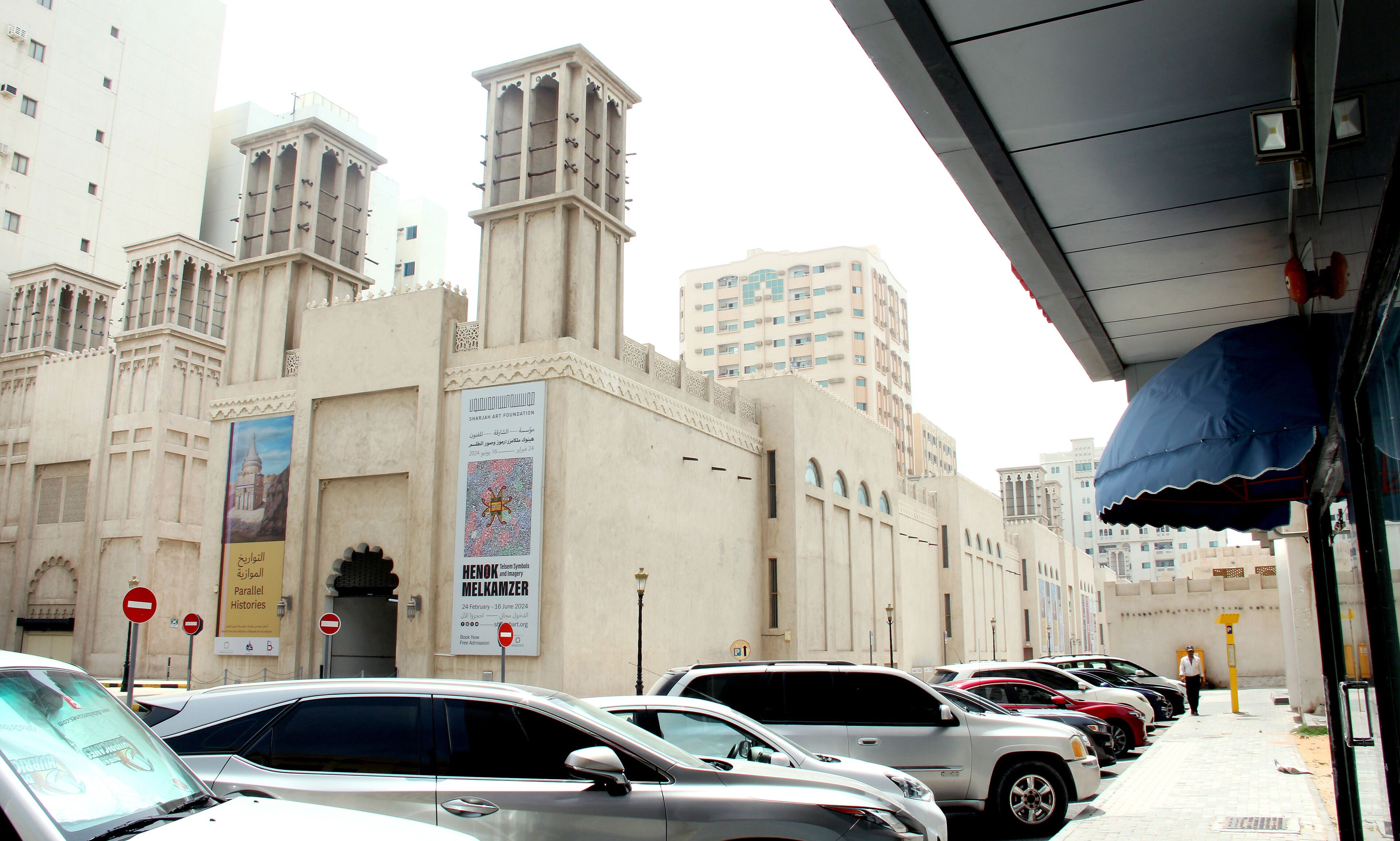Une rangée de voitures garées devant un bâtiment sur lequel est apposée une enseigne indiquant la présence de la Sharjah Art Foundation.