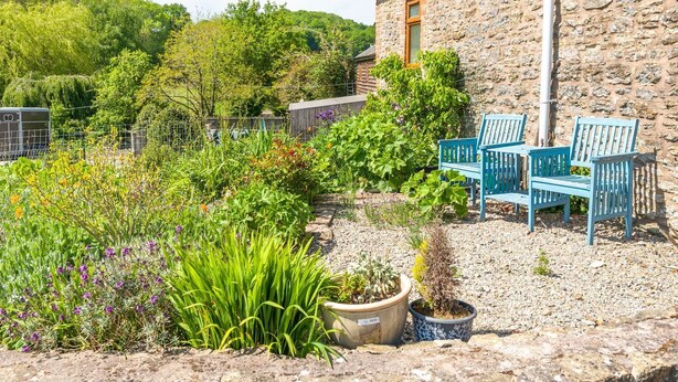 Un edificio de piedra con aspecto rústico, una ventana pequeña y una puerta. En primer plano, hay sillas azules para exteriores y una variedad de plantas en macetas.