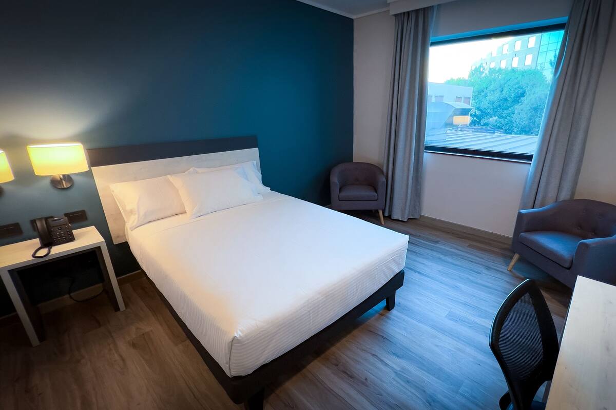 Une chambre d’hôtel équipée d’un lit, d’un bureau, d’une chaise, d’une lampe et d’une fenêtre donnant sur des bâtiments et des arbres.