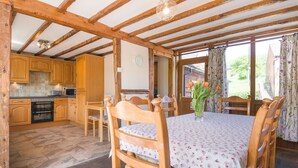 A rustic wooden interior with a dining table set for four, surrounded by wooden chairs. A kitchen with wooden cabinets and a stove is visible in the background. - Owl Barn (Hereford)