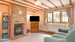 A cozy living room with exposed wooden beams, a stone fireplace, a television, and a window with curtains. - Owl Barn (Hereford)