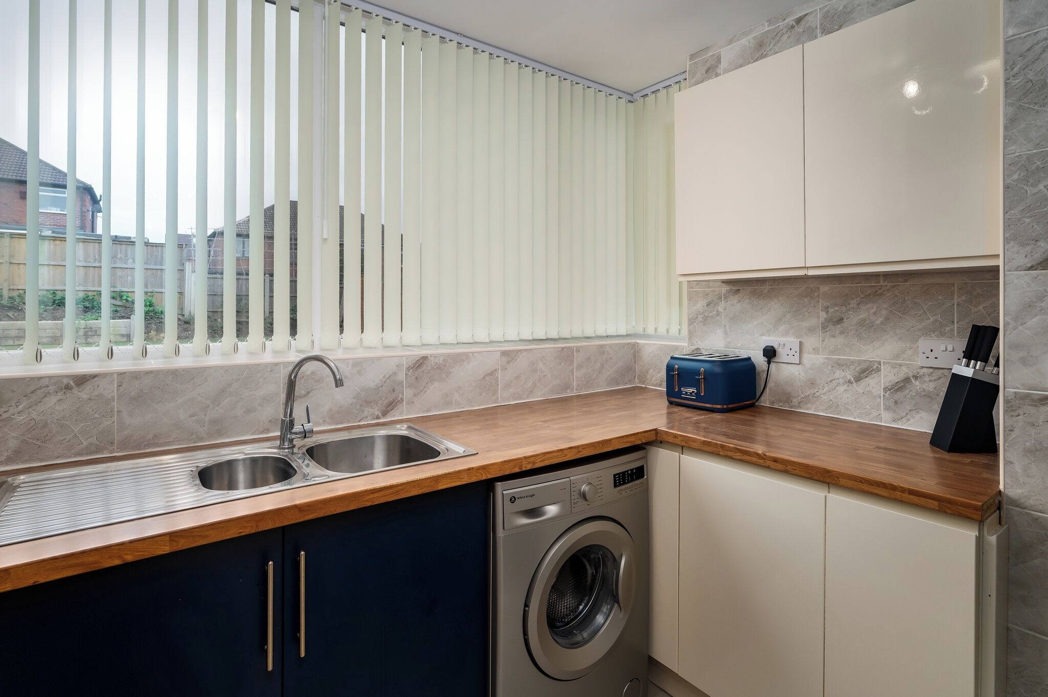 A modern kitchen with a washing machine, a sink, and a window with blinds.