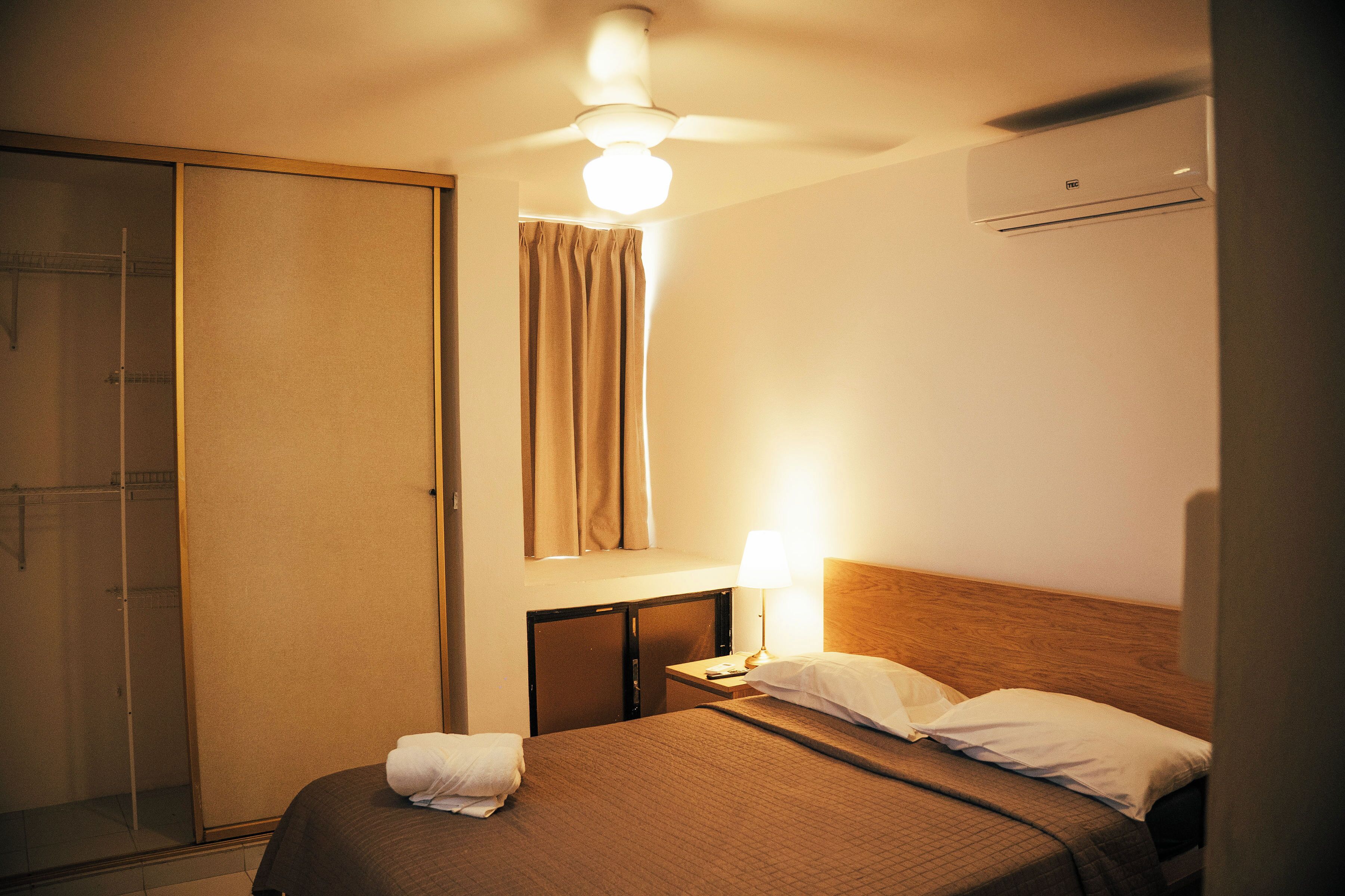 Quarto de hotel com cama, mesa de cabeceira com luminária, guarda-roupa e ventoinha no teto.
