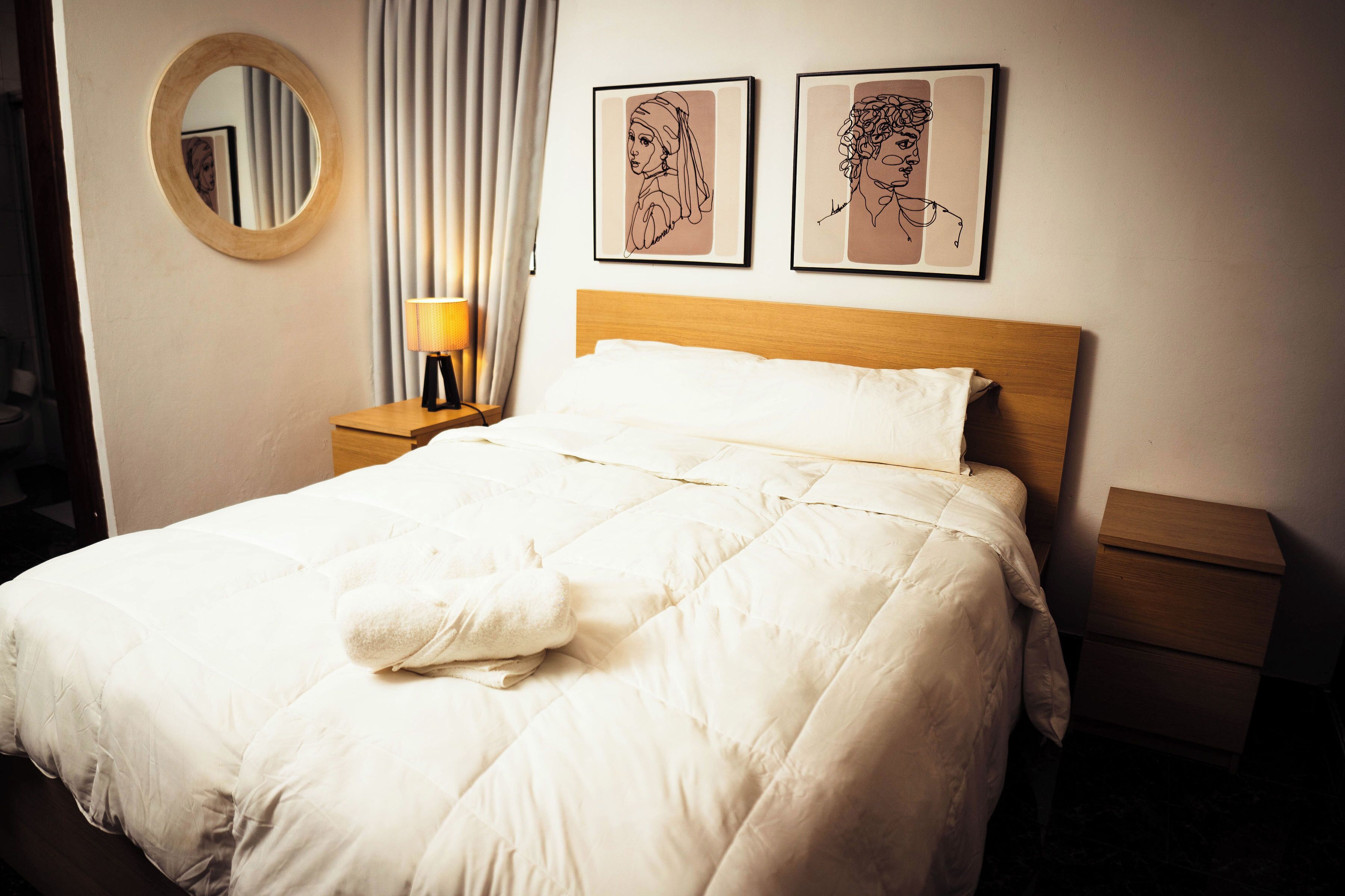 A neatly made bed with a white duvet, a round mirror on the wall, and two framed artworks above it.