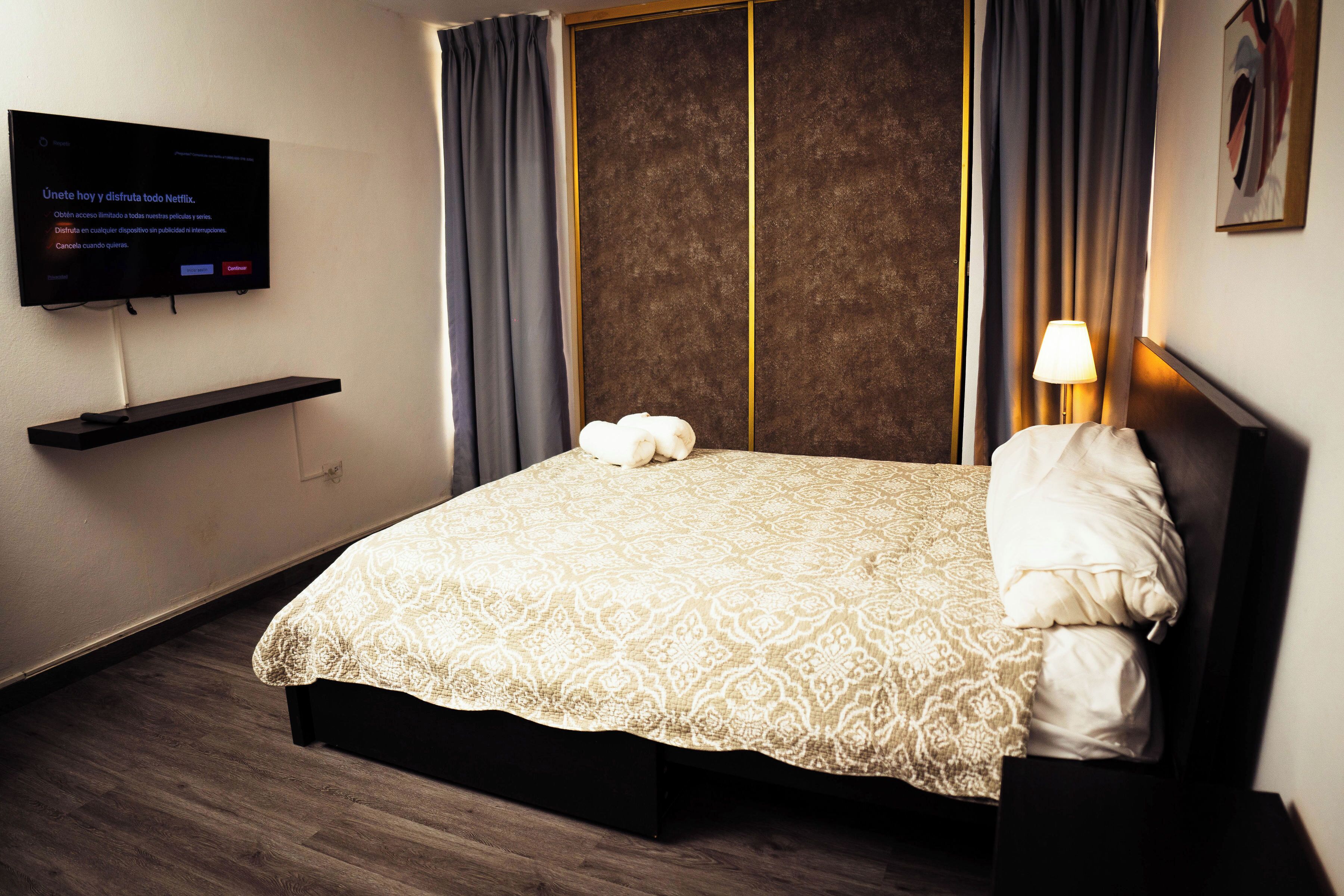 Habitación de hotel con cama, televisor montado en la pared, mesita de noche y lámpara.