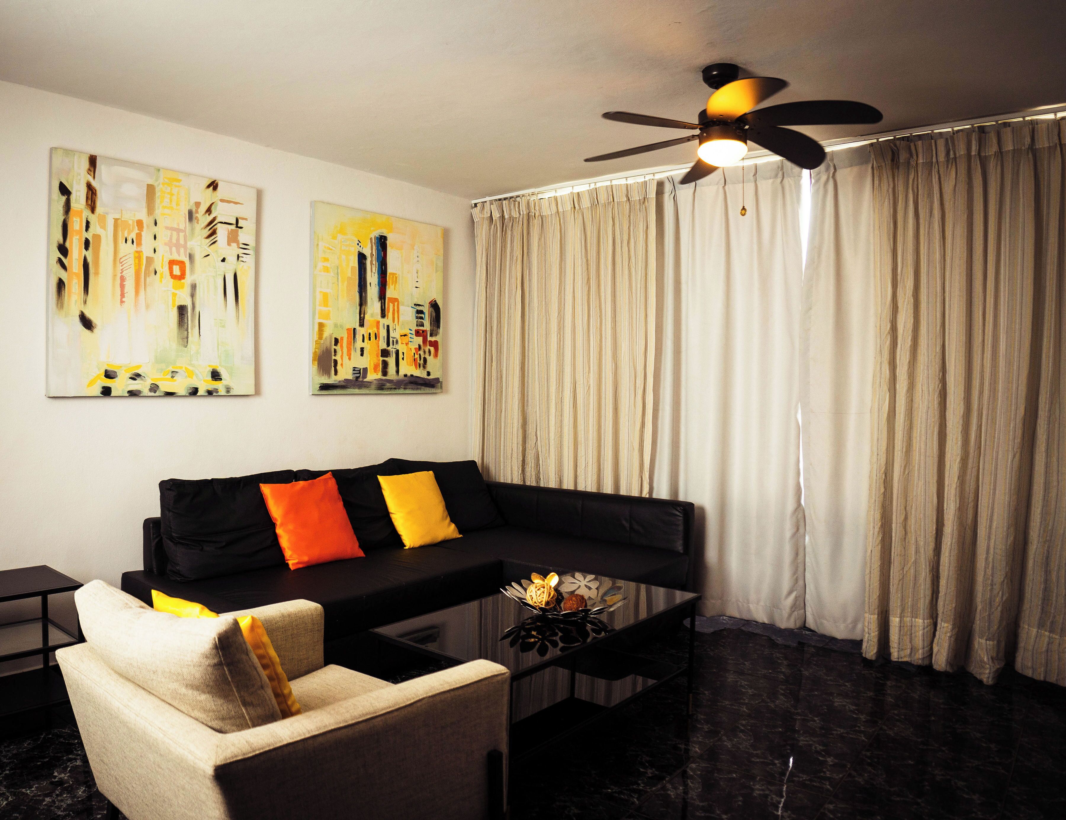 A modern living room with a black sofa, a beige armchair, and a coffee table with decorative items. There are two abstract paintings on the wall and a ceiling fan.