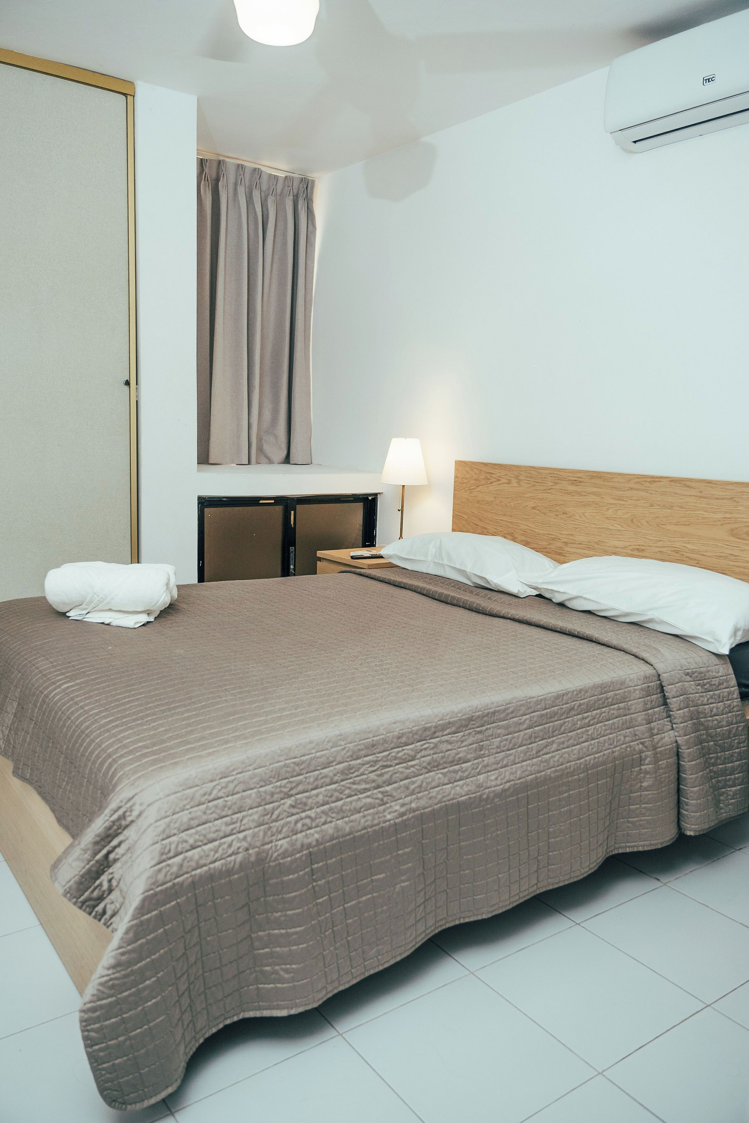 A hotel room with a bed, bedside table, lamp, and an air conditioning unit.