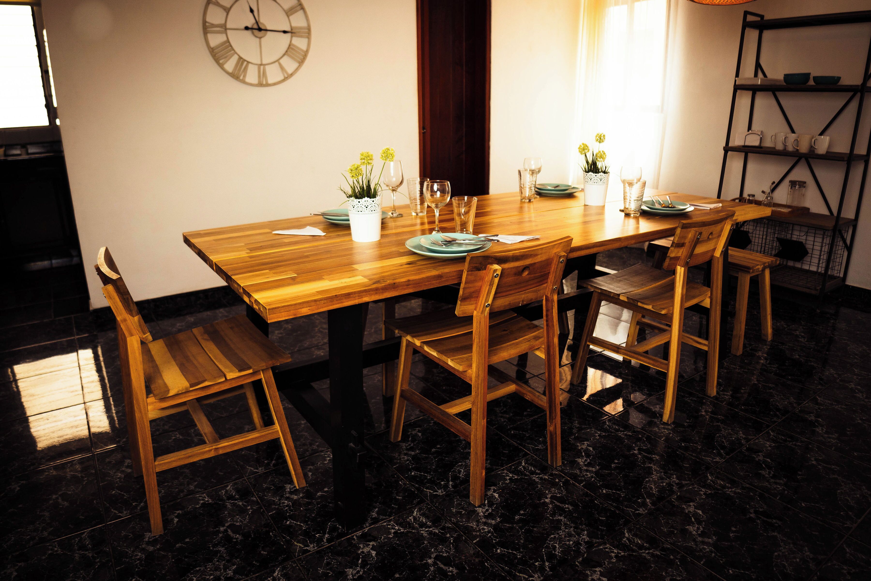 A dining area with a wooden table set for four, surrounded by wooden chairs. A wall clock and a shelf with dishes are visible.