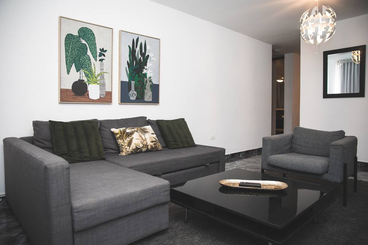A modern living room with a grey sectional sofa, a matching armchair, a black coffee table, and two framed botanical prints on the wall.