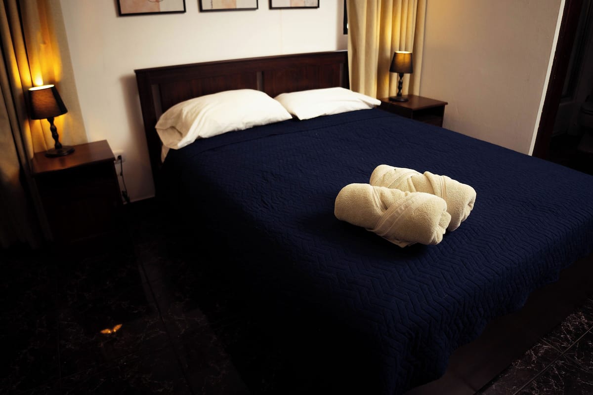 A neatly made bed with a dark blue quilt and two pillows, a pair of white towels on the bed, and two bedside lamps on wooden nightstands.
