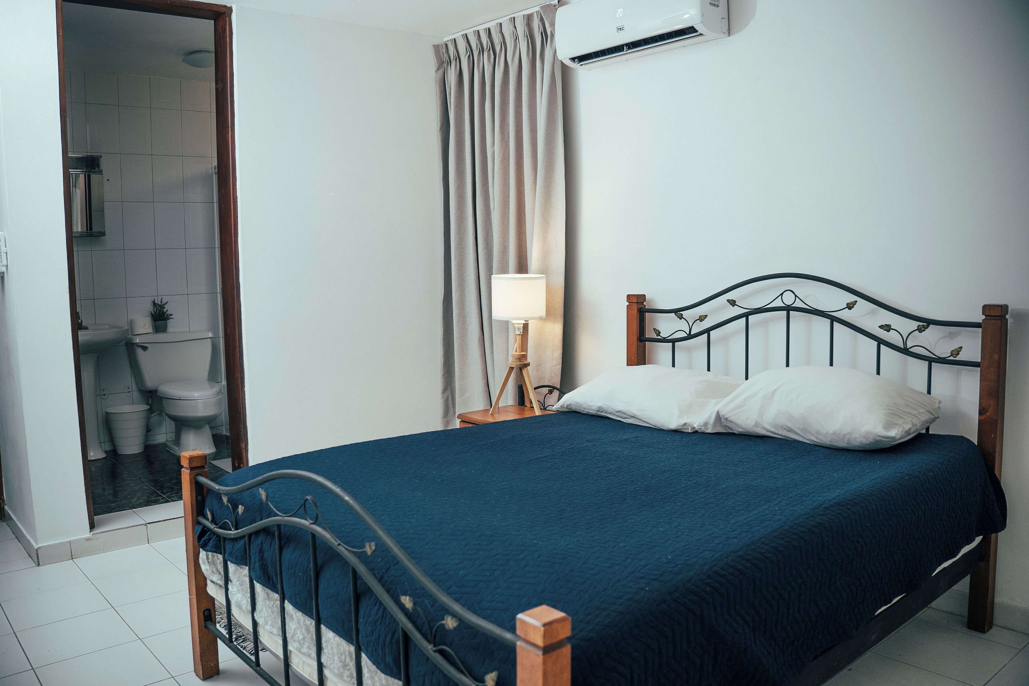 Una cama bien hecha con colcha azul y almohadas blancas, una lámpara de noche y un cabecero de madera.