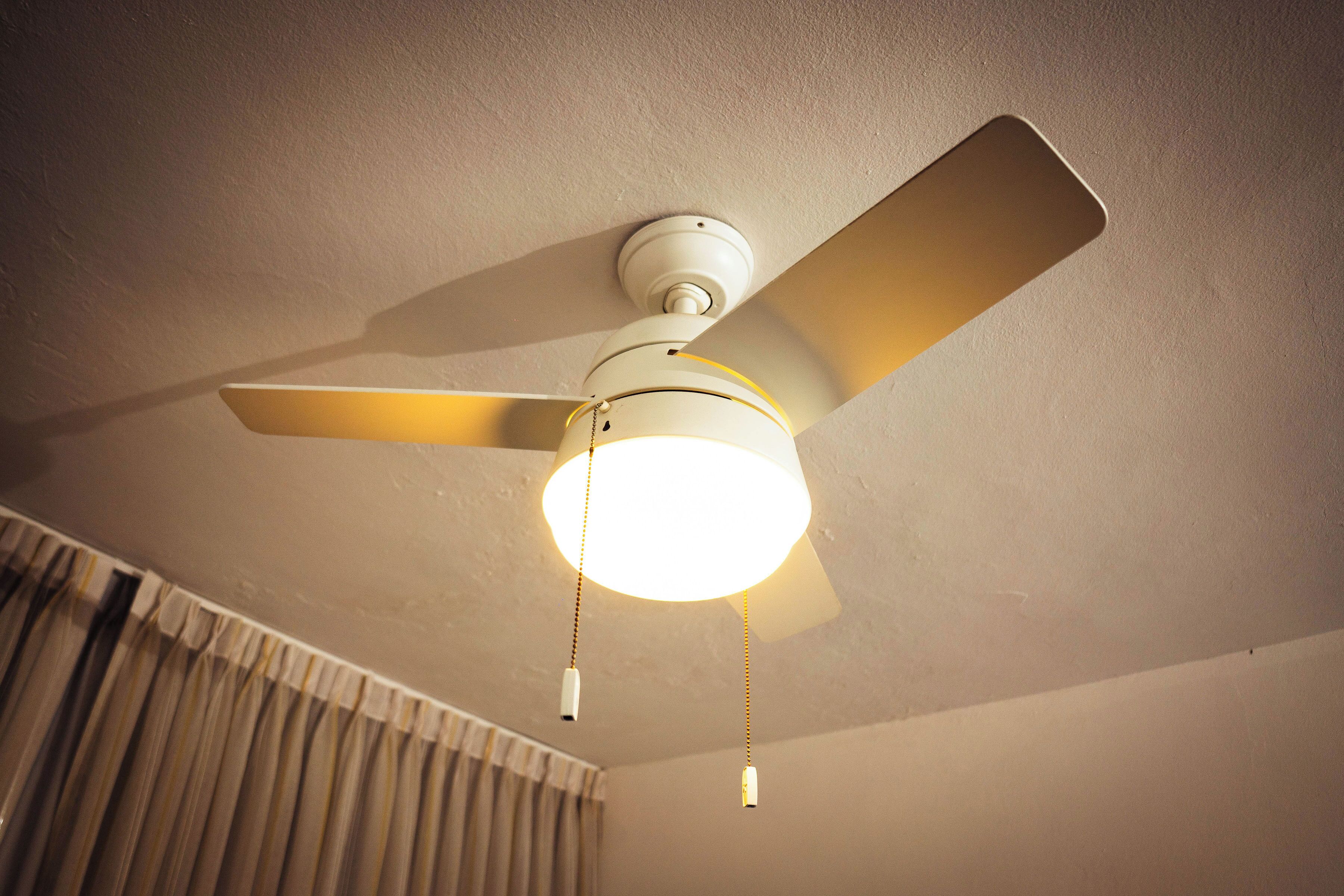 Un ventilateur de plafond avec un luminaire, dans une pièce où se trouve un rideau.