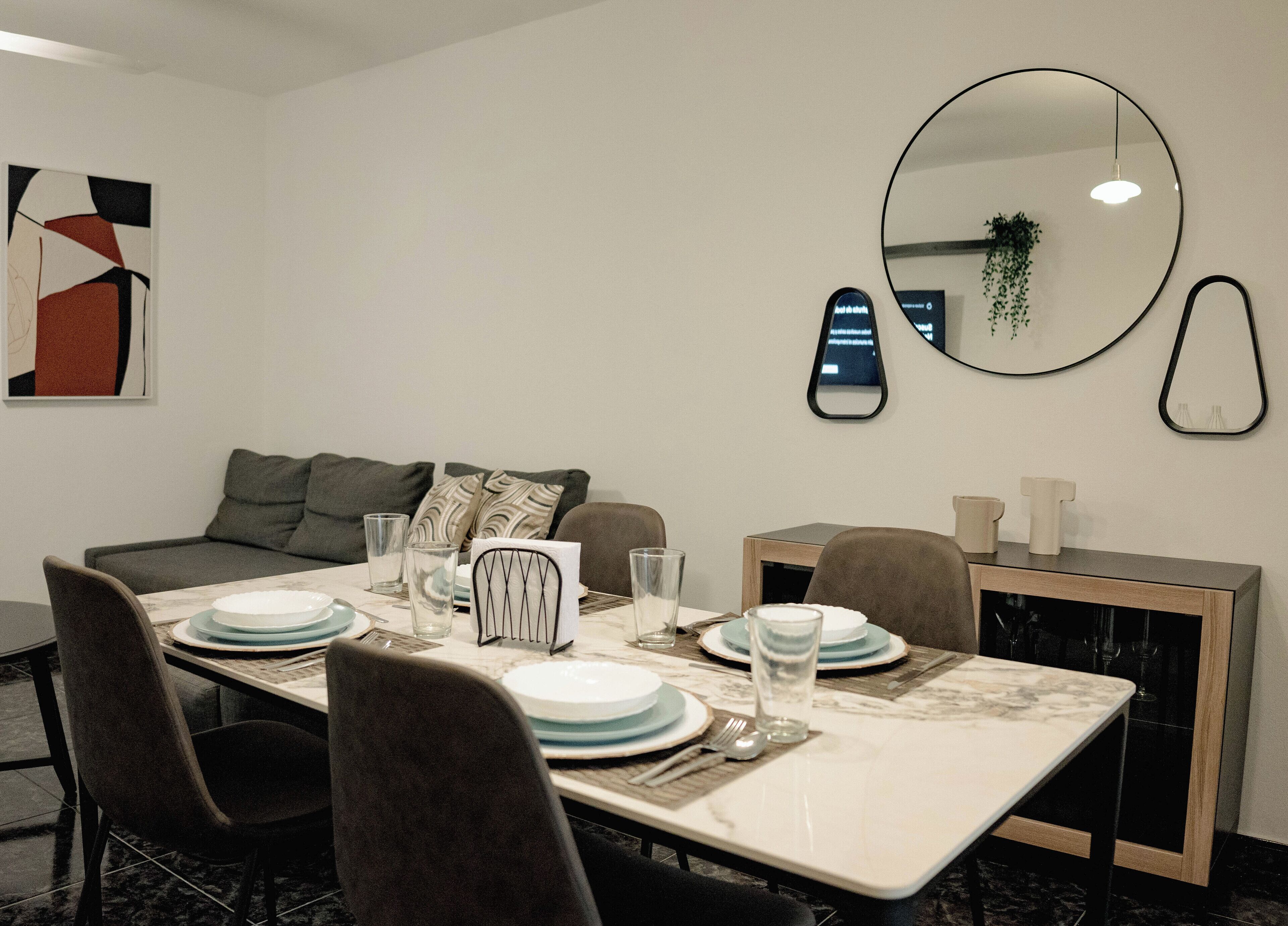 A modern dining area with a marble table, grey upholstered chairs, and a sofa with patterned cushions.