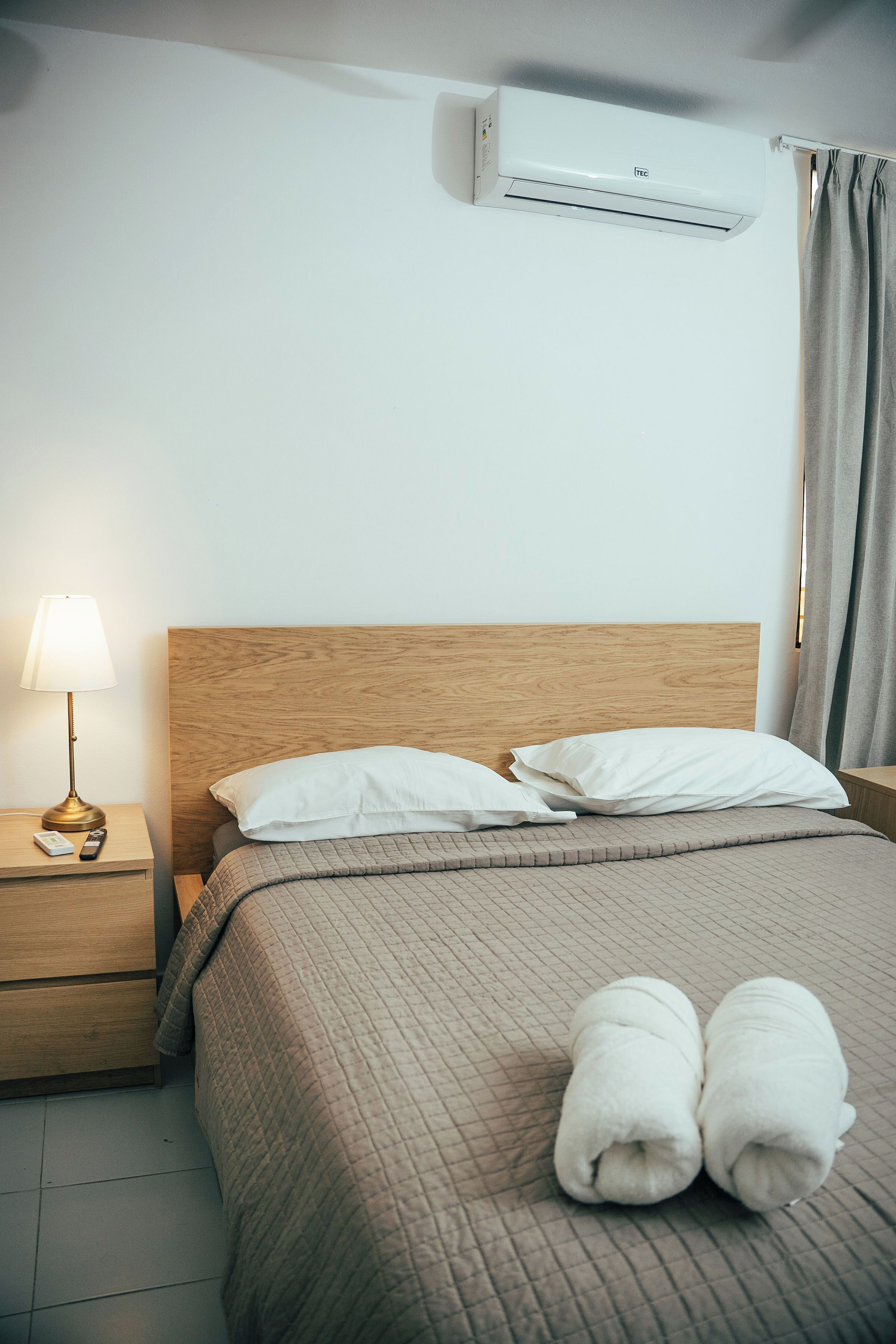 Una cama bien hecha con cabecera de madera, una lámpara de noche y aire acondicionado instalado en la pared.