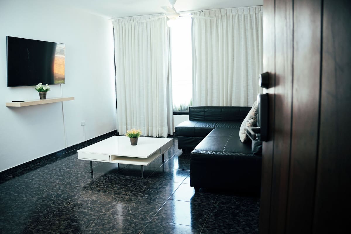 A modern living room with a flat-screen TV, a coffee table, a black leather sofa, and a wooden door.