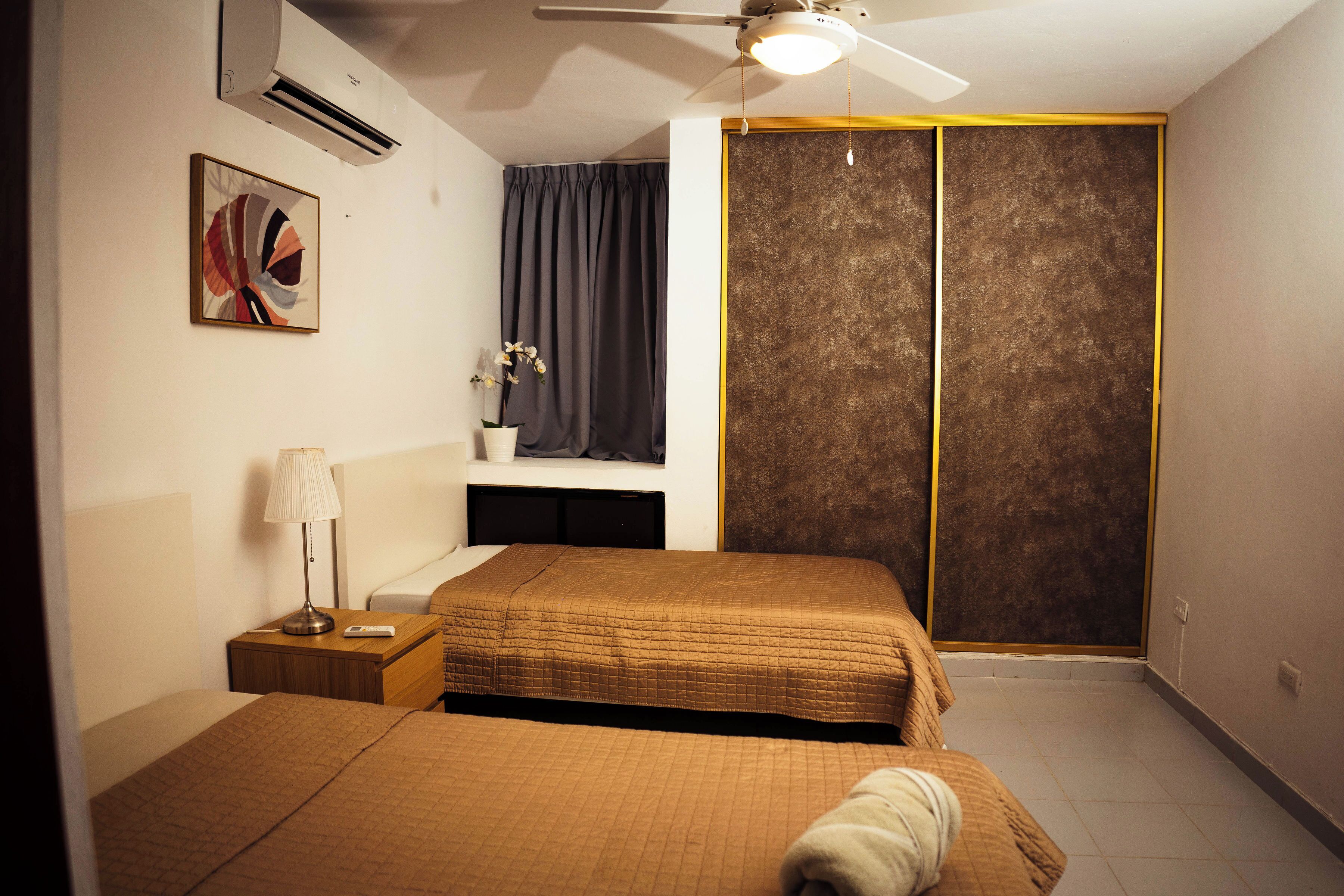 Une chambre d’hôtel avec deux lits, un ventilateur de plafond, un climatiseur, une table de chevet avec une lampe, un petit bureau et une grande porte coulissante.