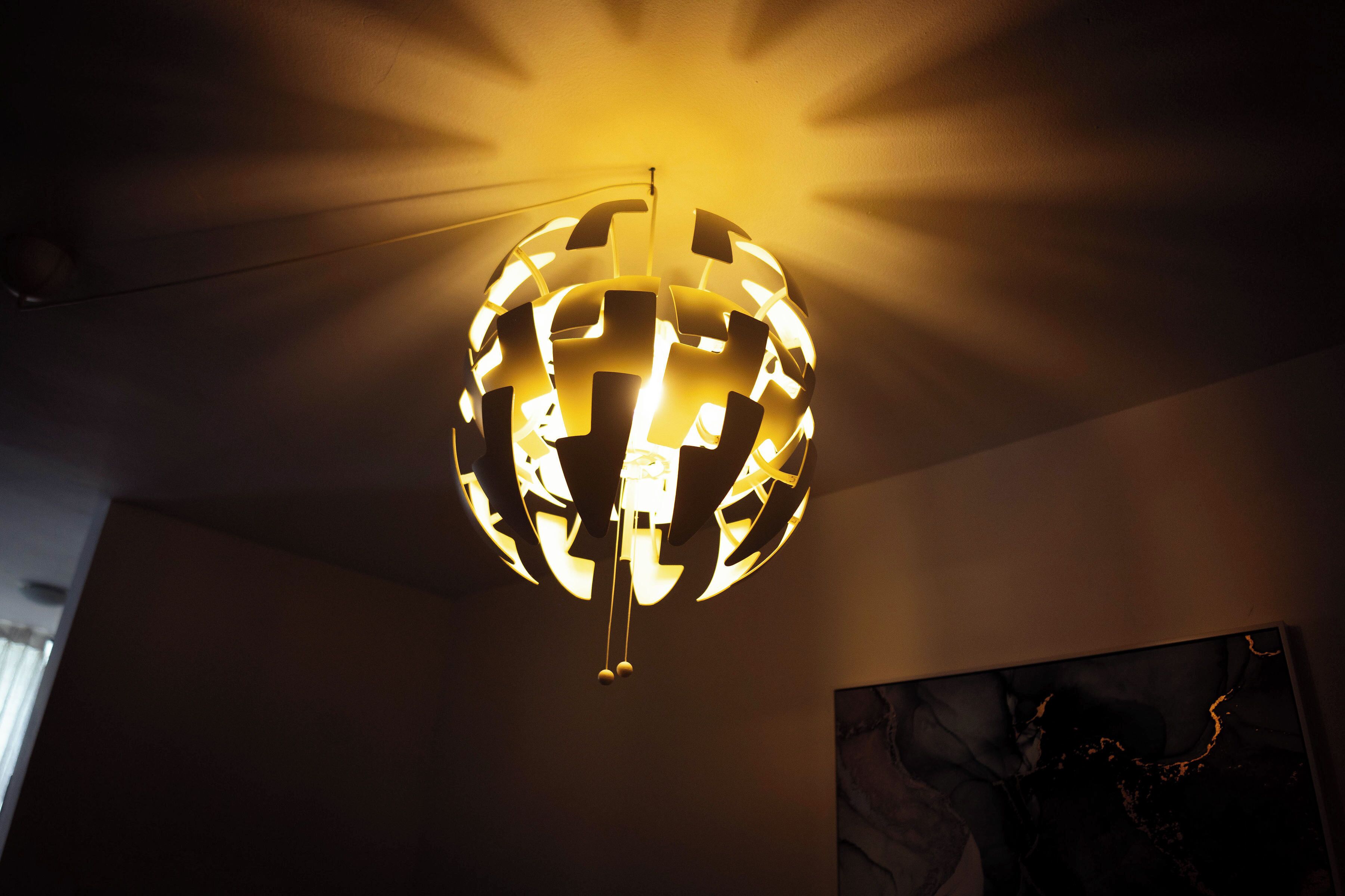 A spherical chandelier with a geometric pattern, hanging from a ceiling in a room with a plain wall and a painting.