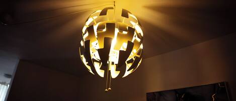 A spherical chandelier with a geometric pattern, hanging from a ceiling in a room with a plain wall and a painting.