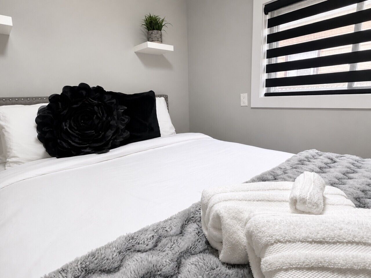 Un lit bien fait, avec une couverture grise et moelleuse, un coussin noir décoratif et une serviette blanche posée sur le côté.
