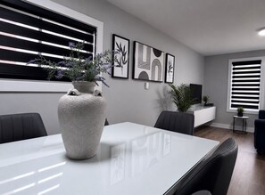 Ein moderner Essbereich mit einem weißen Tisch, schwarzen Stühlen und einer großen Vase mit lila Blumen.