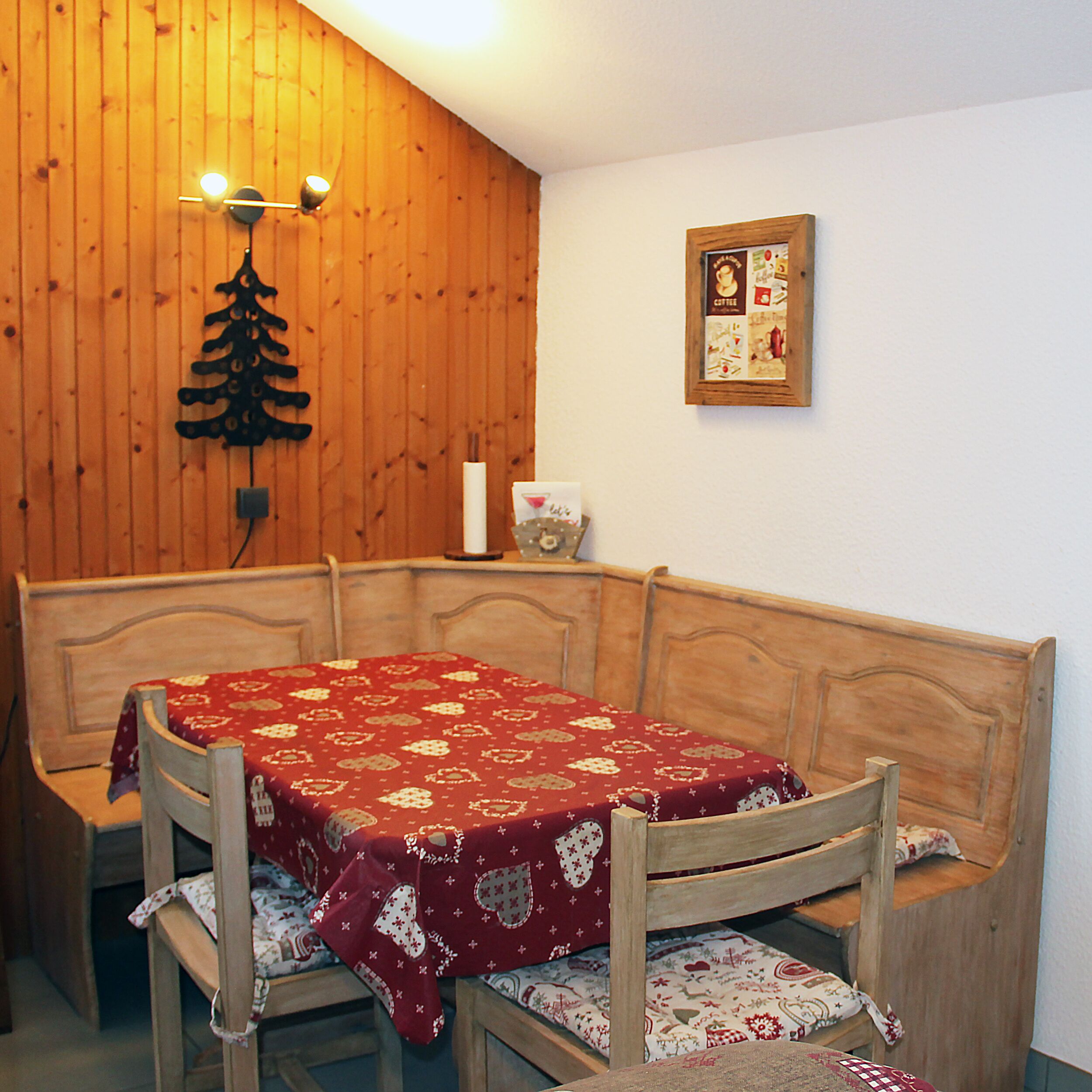 Une table à manger en bois, recouverte d’une nappe rouge, entourée de chaises en bois ornées de coussins à motifs.