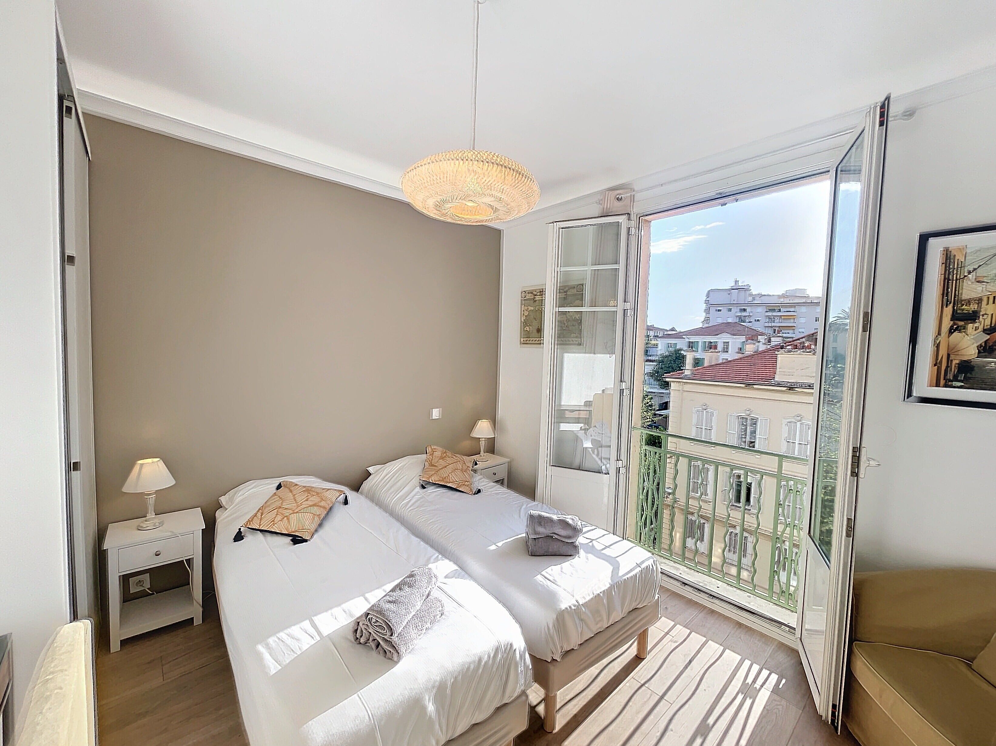 Ein Schlafzimmer mit zwei Betten, einem Nachttisch, einer Lampe, einem Fenster mit Blick auf Gebäude und einem Balkon mit grüner Geländer.