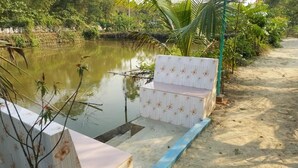 A wooden bench with floral patterns by a calm water body, surrounded by greenery and a dirt path. - Sunapur Mini Resort (Bhurbhuria)