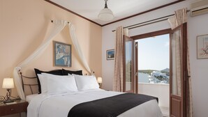 A bedroom with a bed, a nightstand, a window with curtains, and a view of buildings. -  Sunset Paros Naousa (Paros)