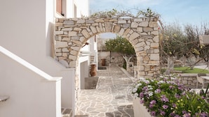 A stone archway leading to a courtyard with a stone floor, surrounded by white walls and potted plants. -  Sunset Paros Naousa (Paros)