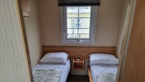 A compact RV bedroom with two single beds, a small wooden nightstand, a window with blinds, and a view of a house outside.