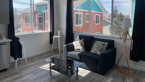 A modern living room with a sofa, a coffee table, and a lamp. There are large windows with curtains, and a view of buildings outside. - Midtown’s Hidden Secret (Reno)
