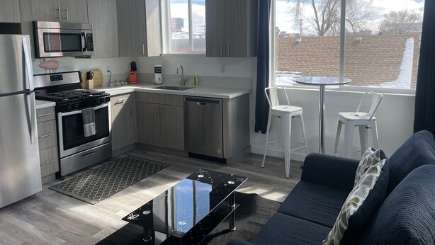 A modern kitchen with stainless steel appliances, a dining area with a round table and two white chairs, and a living room with a dark sofa. - Midtown’s Hidden Secret (Reno)